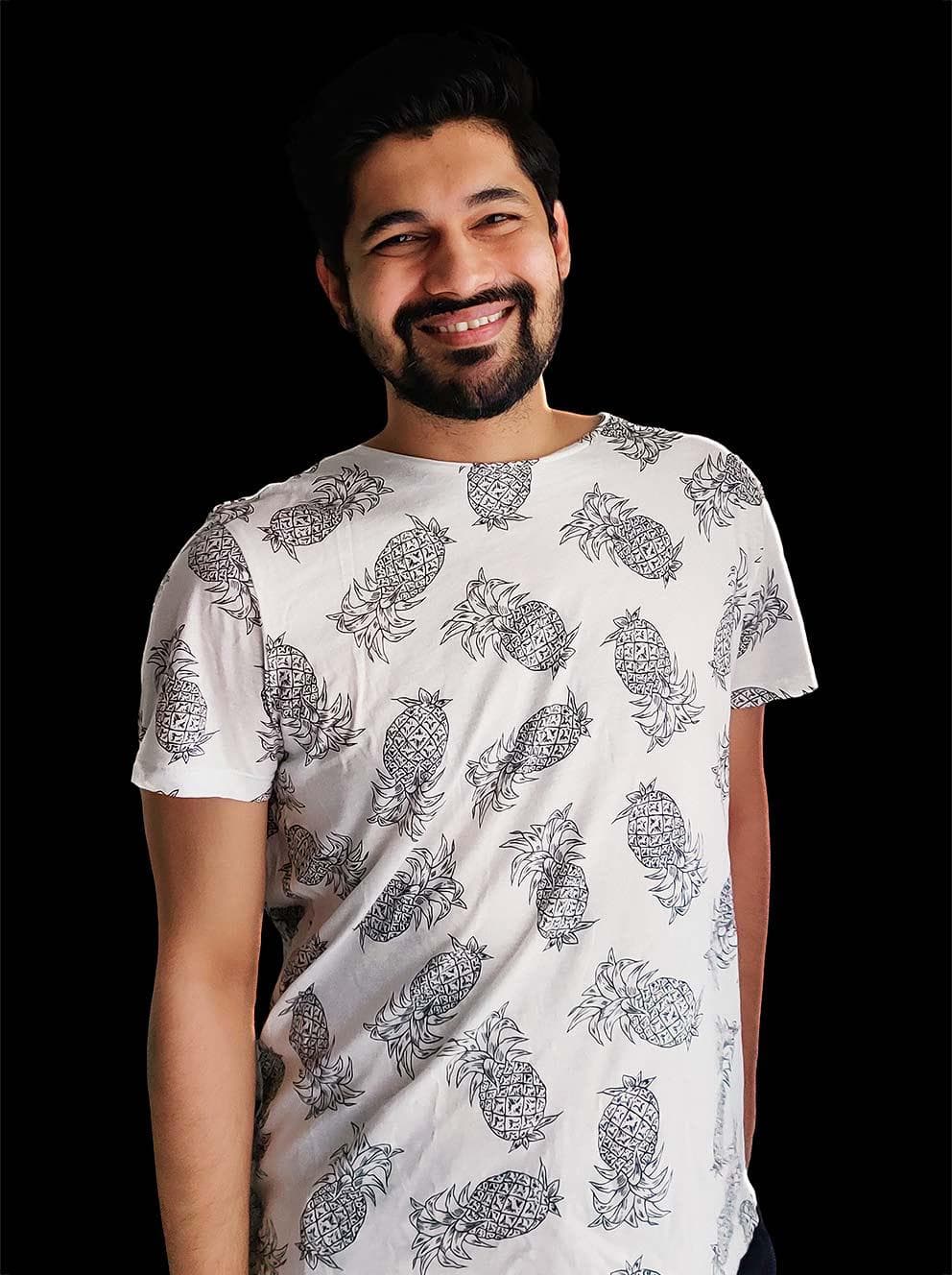 Ganesh Chandrashekar, who is wearing a white t-shirt covered with drawings of black-and-white pineapples, and is standing with his hands at his sides while smiling big
